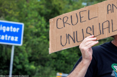 A road sign that says Alligator Alcatraz and an individual in front of it holding a sign that says "Cruel and Unusual."