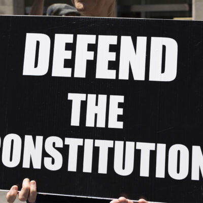 A demonstrator holds a sign (with white letters on a black background) that reads "DEFEND THE CONSTITUTION".