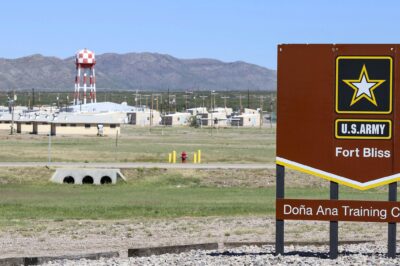 A large brown U.S. Army sign reading “Fort Bliss – Doña Ana Training Complex” stands in the foreground of a wide, open landscape. Behind it are low beige military buildings, utility poles, and a tall red-and-white checkered water tower, with mountains rising in the distance under a clear blue sky.