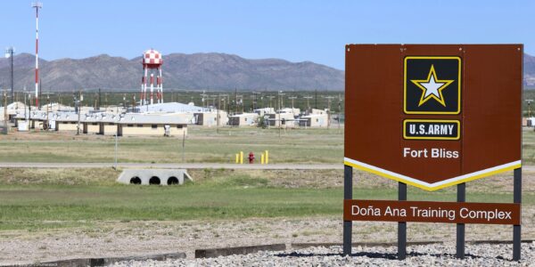A large brown U.S. Army sign reading “Fort Bliss – Doña Ana Training Complex” stands in the foreground of a wide, open landscape. Behind it are low beige military buildings, utility poles, and a tall red-and-white checkered water tower, with mountains rising in the distance under a clear blue sky.