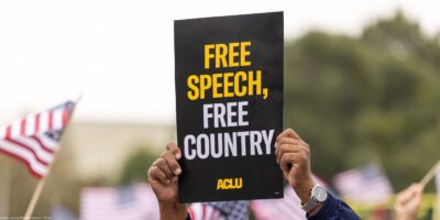 A demonstrator (with only their hands visible) holds up a sign that reads " FREE SPEECH, FREE COUNTRY - ACLU".