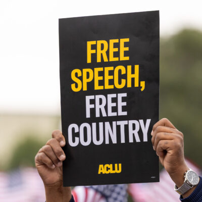 A demonstrator (with only their hands visible) holds up a sign that reads " FREE SPEECH, FREE COUNTRY - ACLU".