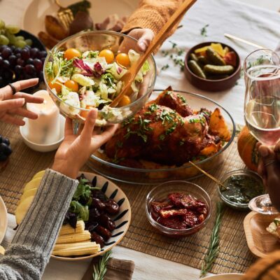 A multi-ethnic group of people sharing a festive, communal meal at a dining table with roasted chicken, salad, bread, fruit, and wine.