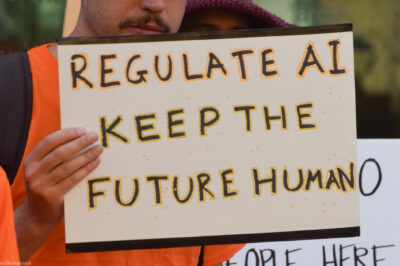 The camera focuses on a sign held by a demonstrator that reads, "REGULATE AI KEEP THE FUTURE HUMAN."