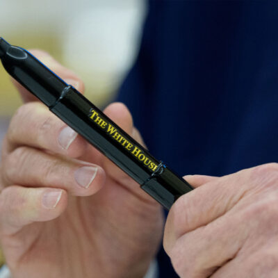A close-up of President Trump’s hands holding a black marker pen inscribed with “The White House” in gold lettering, moments after signing executive orders in the Oval Office.